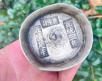 May include: A small, silver-colored metal bowl with Chinese characters stamped inside. The bowl has a slightly irregular shape and a textured surface. The characters are arranged in rectangular blocks. The bowl is held in a hand against a background of green leaves.