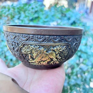 May include: A bronze-colored bowl with intricate relief carvings. The bowl features a detailed design with a golden dragon motif. The rim and base have a darker, contrasting finish, highlighting the detailed artwork. The bowl is held in a hand.
