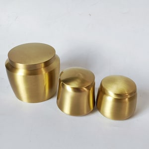 May include: Three brass containers of varying sizes. The containers have a cylindrical shape with rounded lids. The brass has a polished finish, reflecting light. The containers are arranged from largest to smallest.