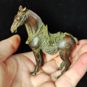 May include: A small, bronze-colored horse figurine with a saddle. The horse has a detailed mane, bridle, and saddle. The figurine is held in a hand, showcasing its size and craftsmanship. The horse is a dark brown color with green accents.
