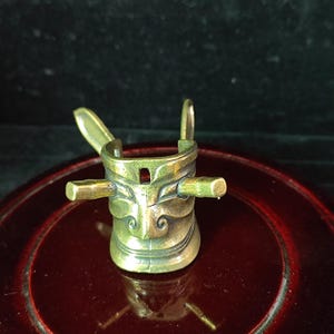 May include: A bronze-colored, antique-style metal object with a unique design. It features a central cup-like structure with a face-like appearance, two horizontal bars, and two vertical loops. The object has an aged, weathered look.