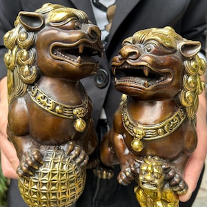 May include: A pair of bronze-toned guardian lion statues with polished gold-coloured accents on the manes, collars, and bells. One lion rests a paw on a textured spherical orb, while the other rests a paw on a smaller, detailed lion cub figure.