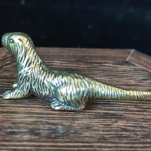 May include: A small, detailed brass figurine of a stylized otter. The sculpture features textured fur and is posed in a reclining position. The figurine is displayed on a wooden surface, showcasing its intricate design and craftsmanship.