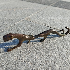 Puede incluir: Escultura estilizada de felino, posiblemente un leopardo o jaguar, en color bronce. La escultura tiene un cuerpo largo y sinuoso y está colocada sobre una superficie gris texturizada. Los detalles incluyen la cabeza, las patas y la cola del animal.