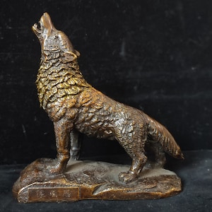 May include: A bronze-colored wolf statue in a howling pose. The wolf stands on a small, textured base. The statue has detailed fur and is set against a dark background. The wolf's head is raised, mouth open in a howl.