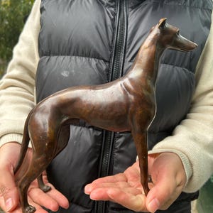 Puede incluir: Escultura de bronce de un perro esbelto, probablemente un galgo, en posición de pie. La escultura tiene un acabado liso y pulido con una pátina marrón oscuro. La forma del perro es alargada, con cuello y patas largas.
