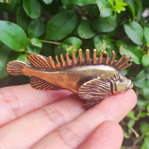 Puede incluir: Una detallada figura de pez de color bronce con aletas texturizadas y una aleta dorsal espinosa. El pez se sostiene contra un fondo de follaje verde, mostrando su intrincado diseño y brillo metálico. La figura es un objeto decorativo.