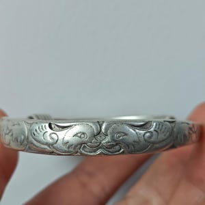 May include: A silver-colored bangle bracelet with detailed carvings. The bracelet features symmetrical animal-like figures and swirling patterns. The metal has a slightly aged appearance, suggesting it may be vintage or antique.