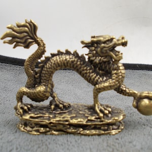 Puede incluir: Figura de dragón de bronce. El dragón está posado sobre una base rocosa, sosteniendo una esfera. El dragón tiene un cuerpo detallado y escamoso, una cola fluida y la boca abierta. La figura es de metal.