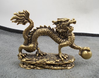 Puede incluir: Figura de dragón de bronce. El dragón está posado sobre una base rocosa, sosteniendo una esfera. El dragón tiene un cuerpo detallado y escamoso, una cola fluida y la boca abierta. La figura es de metal.