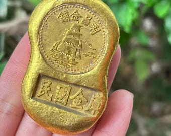 May include: A gold-colored, rectangular coin with rounded edges. The coin features Chinese characters and a detailed engraving of a sailing ship. The coin's surface has a slightly worn appearance, suggesting age or use.