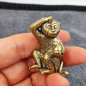 May include: A small, brass-colored monkey figurine. The monkey is in a seated position, with one hand raised to its head and the other holding a small object. The figurine has detailed features and a slightly aged appearance.