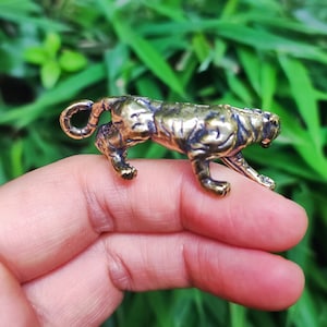 May include: A small, bronze-colored tiger figurine. The tiger is in a walking pose, with its tail curled. The figurine has detailed stripes and a textured surface. The background is blurred green foliage.