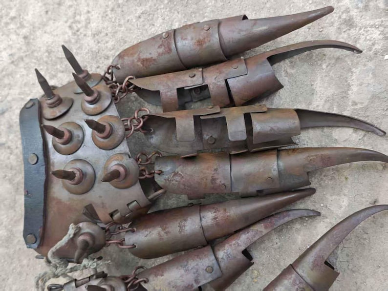 Pu&ograve; includere: Un'arma ad artiglio in metallo arrugginito con lunghe lame ricurve e una protezione per le nocche a punte. L'artiglio &egrave; collegato da catene e presenta un aspetto invecchiato e usurato, che suggerisce un tema storico o fantasy.