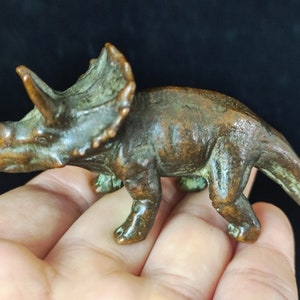 May include: Bronze-colored Triceratops figurine. The dinosaur has three horns, a frill, and four legs. The figurine is a detailed representation of the prehistoric animal. The sculpture is approximately 4 inches long.