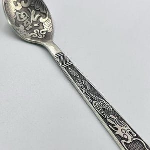 May include: A silver spoon with intricate engraved designs. The spoon's bowl features ornate patterns, while the handle is decorated with a dragon motif. The spoon is made of a shiny, metallic material.