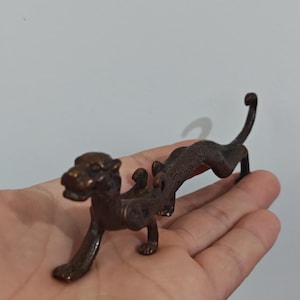 Könnte beinhalten: Eine bronzefarbene Figur einer stilisierten Katzenkreatur mit langgestrecktem Körper und gebogenem Schwanz. Die Skulptur weist detaillierte Merkmale auf, darunter Pfoten, einen Kopf und ein dekoratives Element auf dem Rücken. Die Figur wird in der Hand einer Person vor einem schlichten Hintergrund gehalten.