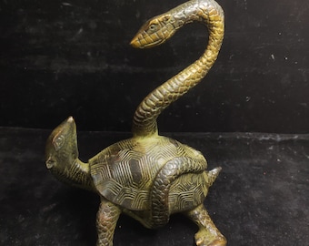 May include: Bronze sculpture of a turtle with a snake wrapped around its shell and neck. The snake's head is raised, and the turtle's shell has a geometric pattern. The sculpture has a weathered, antique appearance.