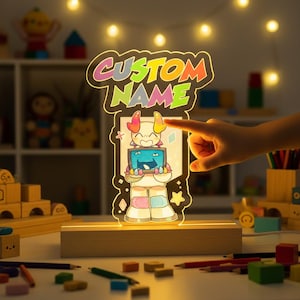 Puede incluir: Luz de noche LED con nombre personalizado con un personaje de dibujos animados sosteniendo una pantalla. La luz tiene una base de madera y las palabras "CUSTOM NAME" en letras coloridas. El personaje tiene cuernos y viste un atuendo rosa y azul.