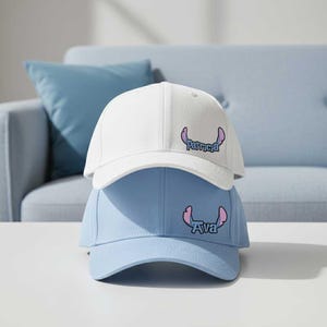 Puede incluir: Dos gorras de béisbol apiladas. La gorra superior es blanca y la inferior azul claro. Cada gorra tiene un gráfico de dibujos animados y un nombre. La gorra blanca dice "Patricia", y la azul claro dice "Ava".