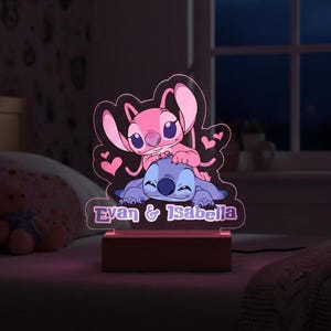 May include: A personalized night light featuring the characters Stitch and Angel from Lilo & Stitch. The pink and blue figures are atop a wooden base, with the names "Evan & Isabella" illuminated below. The light is on a bed.