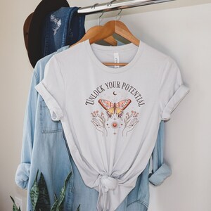 Könnte beinhalten: Ein weißes T-Shirt mit dem Aufdruck "Unlock Your Potential" in Braun. Ein stilisierter Schmetterling mit orangefarbenen und gelben Flügeln ist mittig auf dem Shirt platziert. Der Schmetterling ist von kleinen Blumen und Sternen umgeben.