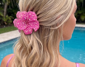 Puede incluir: Una pinza para el cabello en forma de flor de color rosa brillante adornada con numerosos cristales pequeños y brillantes. El accesorio está sujeto en cabello rubio, presentando un diseño floral de cinco pétalos con una base rosa mate y una densa cobertura de pedrería iridiscente.