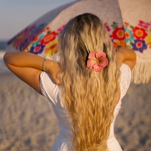 May include: A person with long, wavy blonde hair, styled with a pink flower hair clip, stands on a beach. They wear a white dress and hold a colourful floral parasol. The scene is lit by warm sunlight.