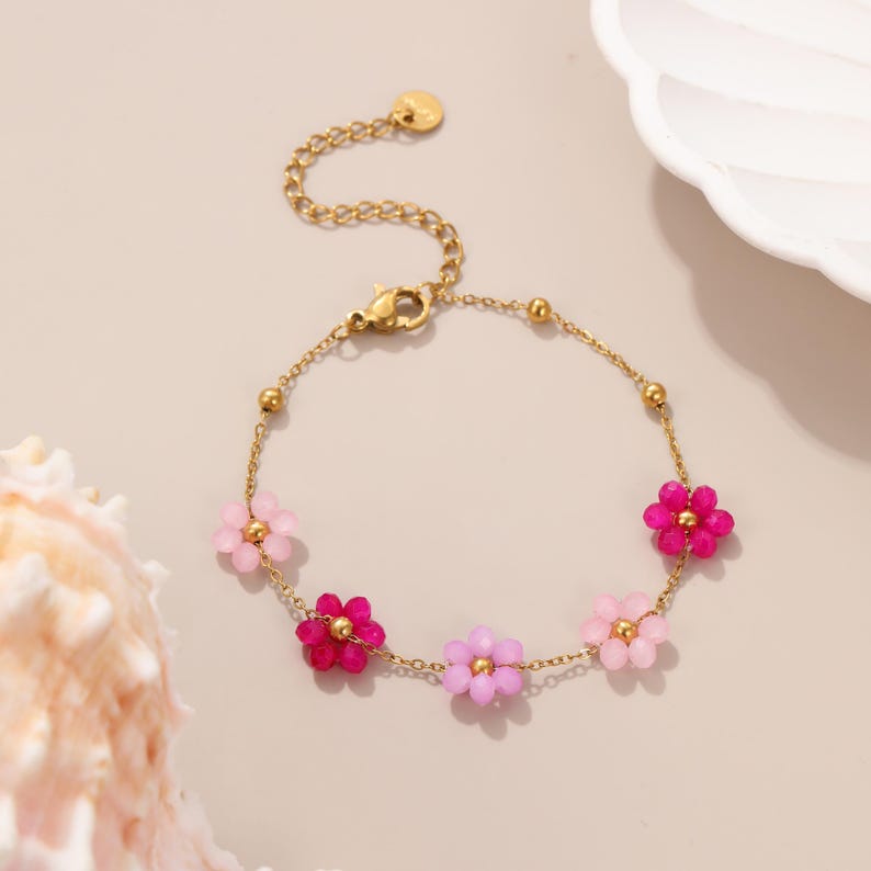 Puede incluir: Pulsera dorada con un delicado dise&ntilde;o de cadena. La pulsera est&aacute; adornada con cinco cuentas en forma de flor en tonos rosa y morado. Tiene un cierre de mosquet&oacute;n y una cadena ajustable. Aproximadamente 18 cm de largo.