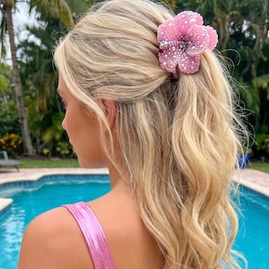 Puede incluir: Una pinza para el cabello con forma de flor rosa adornada con numerosas cuentas pequeñas y cristales brillantes. El accesorio está sujeto en cabello rubio, presentando un degradado de tonos rosa claro a rosa intenso en sus pétalos.