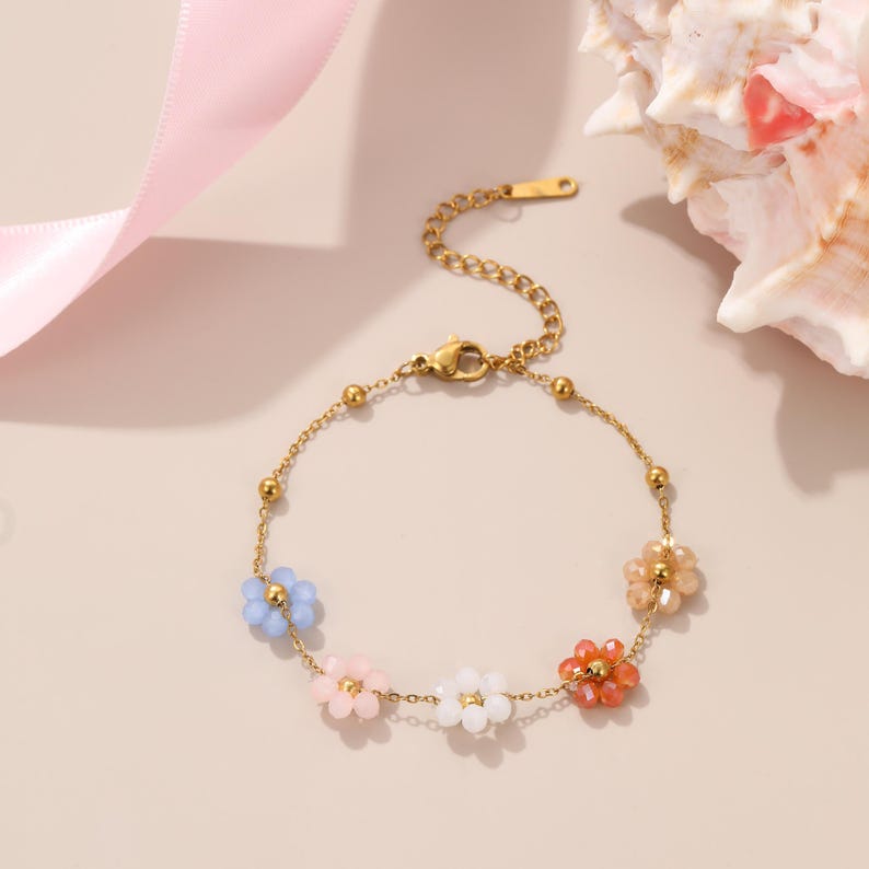 Puede incluir: Una pulsera dorada con una delicada cadena y coloridos abalorios de flores con cuentas. La pulsera tiene un cierre de mosquet&oacute;n y est&aacute; adornada con detalles florales azules, rosas, blancos y naranjas. Una joya encantadora.