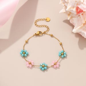 Puede incluir: Pulsera dorada con una delicada cadena y cinco colgantes de flores. Las flores alternan entre azul claro y rosa, con cuentas doradas en el centro. La pulsera tiene una longitud ajustable y un cierre de mosquet&oacute;n.