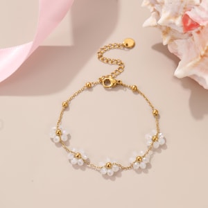 Puede incluir: Pulsera dorada con una delicada cadena y peque&ntilde;os abalorios de flores blancas. La pulsera tiene un cierre de mosquet&oacute;n y una cadena ajustable. La pulsera se muestra sobre una superficie rosa claro con una concha marina en el fondo.
