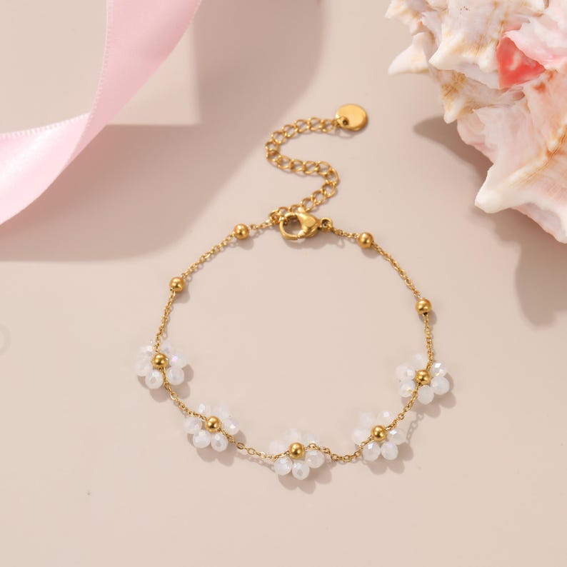 Puede incluir: Pulsera dorada con una delicada cadena y peque&ntilde;os abalorios de flores blancas. La pulsera tiene un cierre de mosquet&oacute;n y una cadena ajustable. La pulsera se muestra sobre una superficie rosa claro con una concha marina en el fondo.