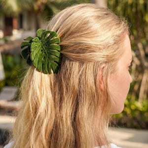 Puede incluir: Una pinza para el cabello de terciopelo verde oscuro con forma de hoja de monstera. El accesorio presenta una superficie texturizada suave al tacto con venas de hoja detalladas y un diseño de varias capas, sujetando un mechón de cabello rubio.