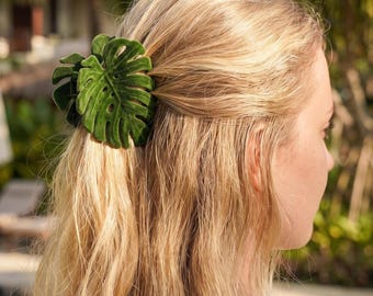 Puede incluir: Una pinza para el cabello de terciopelo verde oscuro con forma de hoja de monstera. El accesorio presenta una superficie texturizada suave al tacto con venas de hoja detalladas y un diseño de varias capas, sujetando un mechón de cabello rubio.