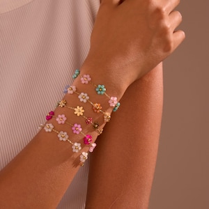 Puede incluir: Varios delicados brazaletes dorados adornados con coloridos diseños florales de cuentas se muestran en una muñeca. Las flores son en tonos de rosa, azul, blanco y amarillo. Los brazaletes están unidos por una fina cadena de oro.
