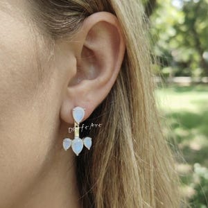 Pode incluir: Brincos de prata com um conjunto de pedras preciosas iridescentes em forma de pêra. Os brincos são projetados para serem usados no lóbulo da orelha e se estendem para baixo com três pedras preciosas adicionais.