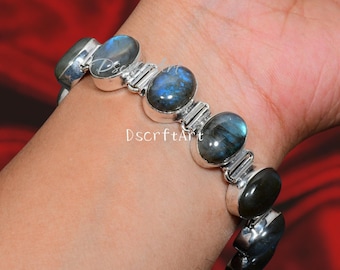 May include: A silver bracelet featuring oval-shaped labradorite stones. The stones display iridescent blue and green hues. The bracelet is connected by silver links and is worn on a wrist. The text "DscrftArt" is visible.
