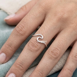 Può includere: Un anello color argento con un design a onda. L'anello è indossato su un dito. L'anello è in stile minimalista ed è probabilmente realizzato in argento sterling. L'anello è un gioiello.