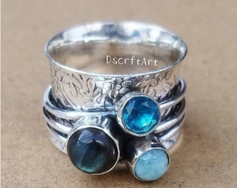 Puede incluir: Un anillo giratorio plateado con tres piedras preciosas. El anillo presenta un patrón floral en la banda exterior y tres piedras preciosas redondas en tonos azules y verdes. El texto "DscrftArt" es visible en el anillo.