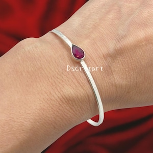 Könnte beinhalten: Ein silberfarbenes Armreif-Armband mit strukturiertem Band und einem tropfenförmigen, tiefroten Edelstein. Das offene Armband wird am Handgelenk getragen, vor rotem Hintergrund.