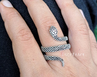 Puede incluir: Un anillo plateado con forma de serpiente se enrolla alrededor de un dedo. La cabeza de la serpiente tiene un patrón texturizado, y el cuerpo se enrolla alrededor del dedo. Una joya única.