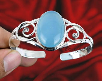 May include: Silver-toned cuff bracelet featuring a large, oval, light blue gemstone. The bracelet has an open design with decorative swirls on either side of the gemstone. The background is a deep red.