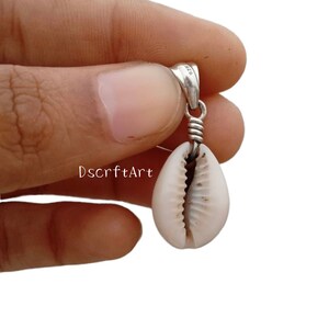 Puede incluir: Un colgante de plata con una concha blanca natural. La concha cuelga de una fianza de plata con un detalle de alambre enrollado. El colgante se sostiene en una mano sobre un fondo blanco. El texto "DscrftArt" es visible.