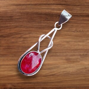 May include: A silver pendant with a large, oval-shaped, red gemstone. The pendant has a unique design with a loop at the top for a chain and a decorative silver frame around the gemstone. The background is a wooden surface.