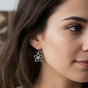 May include: A close-up of a silver flower-shaped earring. The earring has an open design with five petals. It hangs from a silver hook. The image shows a person wearing the earring.