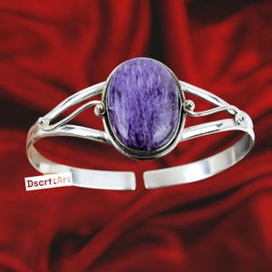 Pode incluir: Uma pulseira de punho prateada com uma pedra preciosa oval roxa. A pedra preciosa é colocada em uma moldura prateada. A pulseira tem um design aberto. O fundo é vermelho escuro.