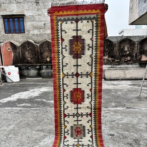 May include: A long, rectangular rug with a cream-coloured central field and a geometric pattern. The border is red, yellow, and grey. The rug has a fringed edge and is held up against a concrete background.
