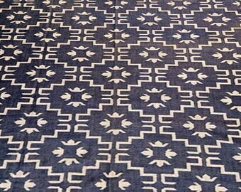 May include: A navy blue and cream-colored patterned rug with a geometric design. The rug is rectangular and lies on a wooden floor. A wooden bench with a white cover and a wooden cabinet are in the background.